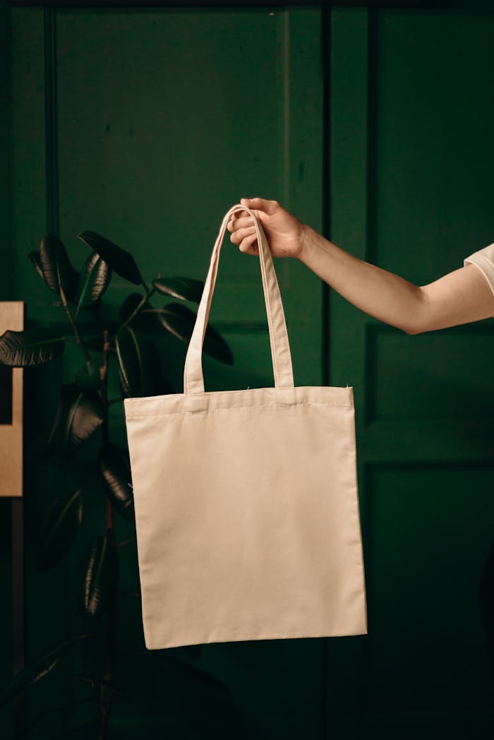 about-01 Simple cotton tote bag held by a hand against a green background, ideal for eco themes.